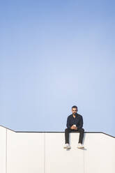 Mann benutzt Telefon, während er auf einem Bauwerk vor blauem Himmel sitzt - MASF18361