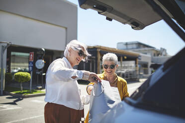 Ältere Freundinnen laden Einkaufstaschen auf einem sonnigen Parkplatz in den Kofferraum eines Autos - CAIF27389