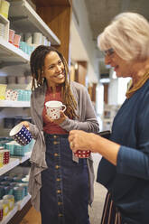 Smiling worker helping senior woman shopping for mugs - CAIF27444