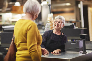 Glückliche ältere weibliche Kassiererin, die Kunden an der Supermarktkasse begrüßt - CAIF27452
