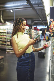 Lächelnde Frau mit Smartphone beim Lebensmitteleinkauf im Supermarkt - CAIF27468