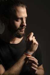 Portrait of bearded young man wearing rainbow wristband against dark background - EGAF00059