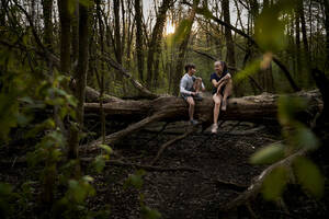 Lächelnde Geschwister in voller Länge im Gespräch auf einem umgestürzten Baum im Wald sitzend - AUF00391