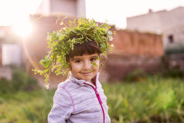 Porträt eines niedlichen Mädchens, das eine Pflanzenkrone trägt, während es im Obstgarten steht - GEMF03678