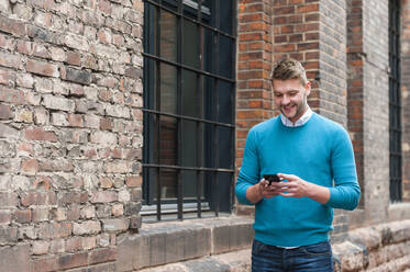 Lächelnder junger Geschäftsmann mit Smartphone an einem Backsteingebäude - DIGF10427