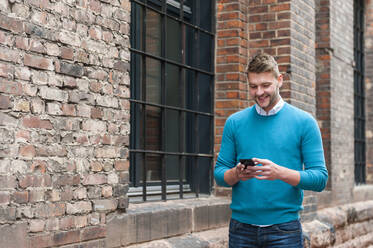 Smiling young businessman using smartphone at a brick building - DIGF10427