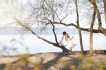 Mid adult couple kissing by bare tree at lakeshore - DIGF10454