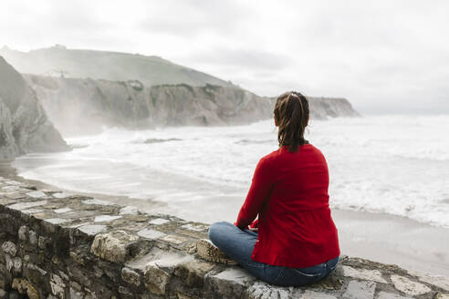 Rückansicht in voller Länge eines entspannten Touristen, der auf einer Stützmauer sitzt und die Klippen gegen den Himmel betrachtet, Itzurun, Zumaia, spanisches Baskenland, Spanien - XLGF00140