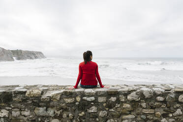 Rear view of relaxed female tourist sitting on retaining wall while looking at sea against sky, Itzurun, Zumaia, Spanish Basque Country, Spain - XLGF00141