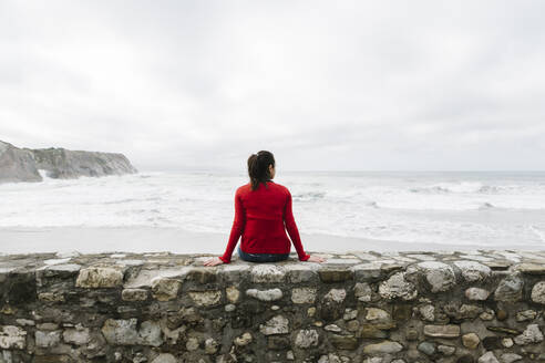 Rückansicht einer entspannten Touristin, die auf einer Stützmauer sitzt und das Meer gegen den Himmel betrachtet, Itzurun, Zumaia, spanisches Baskenland, Spanien - XLGF00141