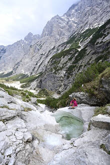 Ältere weibliche Touristin sitzt auf einem Felsen am Bach vor den Bergen - ECPF00908