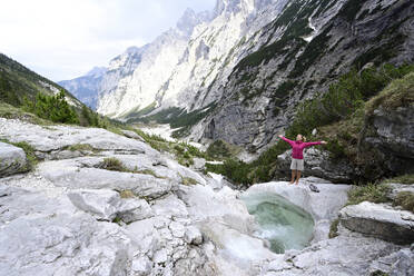 Female tourist standing with arms outstretched on rock by stream against mountains - ECPF00914