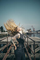 Junge tätowierte Frau wirft ihr langes blondes Haar, während sie auf dem Pier gegen den klaren Himmel an einem sonnigen Tag steht - MTBF00389