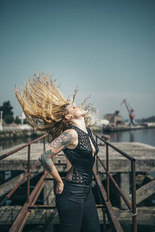 Young tattooed woman tossing her long blond hair while standing on pier against clear sky during sunny day - MTBF00389