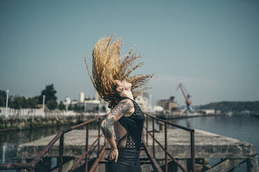 Side view of young tattooed woman tossing her long blond hair while standing hands in pockets on pier during sunny day - MTBF00390