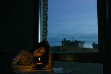 Lonely birthday woman looking at burning candle on cupcake by window in room during sunset - GMLF00183