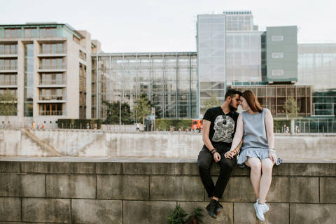 Junges Paar sitzt auf einer Mauer am Flussufer, Berlin, Deutschland, lizenzfreies Stockfoto
