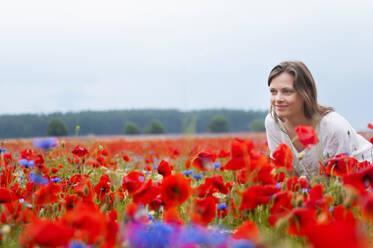 Lächelnde schöne Frau inmitten frischer roter Mohnblumen, die auf einem Feld gegen den klaren Himmel blühen - DIGF10469