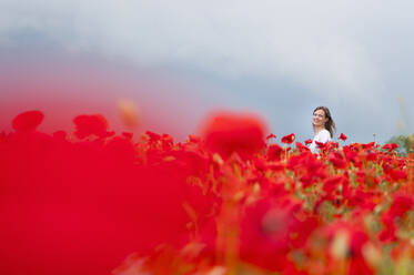 Smiling beautiful woman enjoying on red poppy field against sky - DIGF10477