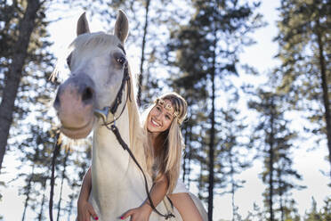 Niedriger Blickwinkel der lächelnden jungen Frau lehnt auf weißem Pferd im Wald - SNF00104
