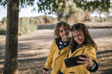 Lächelndes Mädchen nimmt Selfie mit Bruder von Smartphone im Freien - GRCF00203