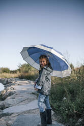 Lächelndes Mädchen mit Regenmantel in voller Länge und mit Regenschirm vor blauem Himmel stehend - GRCF00205