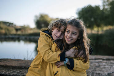 Boy hugging smiling sister wearing yellow raincoats at riverbank - GRCF00208