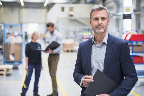 Porträt eines selbstbewussten reifen Geschäftsmannes in einer Fabrik mit Kollegen im Hintergrund - DIGF10500