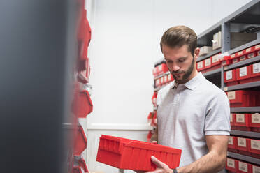 Junger Mann hält einen Karton an einem Regal in einer Fabrik - DIGF10569