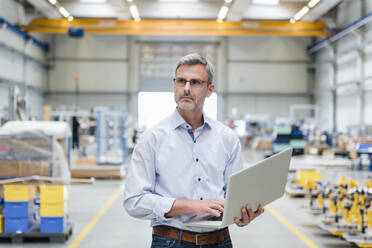 Mature man holding laptop on factory shop floor - DIGF10634