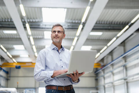 Älterer Mann hält Laptop in einer Fabrikhalle, lizenzfreies Stockfoto