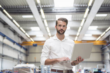 Young man holding laptop on factory shop floor - DIGF10637