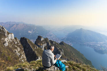 Rear view of hiker reading a book on mountaintop, Orobie Alps, Lecco, Italy - MCVF00313