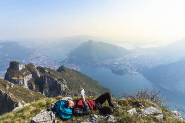 Rear view of hiker reading a book on mountaintop, Orobie Alps, Lecco, Italy - MCVF00314