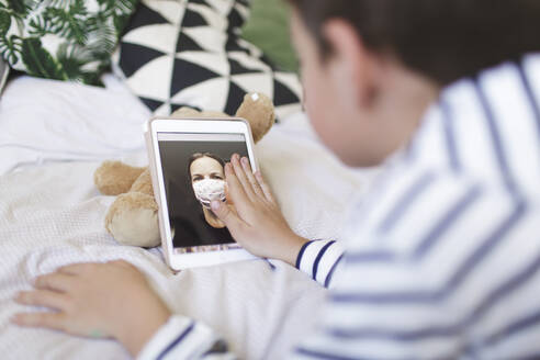 Auf dem Bett liegender Junge, der mit seiner Mutter, die eine Schutzmaske trägt, ein Videogespräch auf einem digitalen Tablet führt - HMEF00925