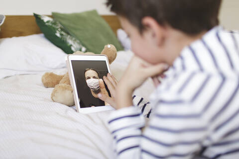 Junge auf dem Bett liegend, der mit seiner Mutter, die eine Schutzmaske trägt, einen Videogespräch auf einem digitalen Tablet führt, lizenzfreies Stockfoto