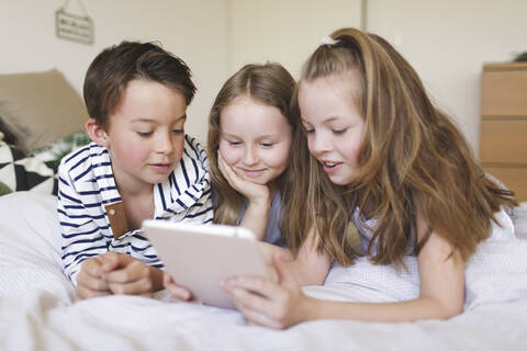 Porträt eines Jungen und seiner beiden Schwestern, die auf einem Bett liegen und ein digitales Tablet für den Hausunterricht benutzen, lizenzfreies Stockfoto