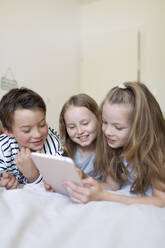 Porträt eines Jungen und seiner beiden Schwestern, die auf einem Bett liegen und ein digitales Tablet für den Hausunterricht benutzen - HMEF00929