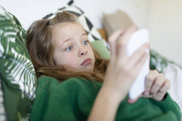 Porträt eines Mädchens mit grünem Pullover, das auf dem Bett liegt und auf sein Handy starrt - HMEF00933