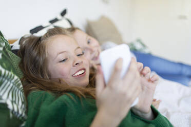 Portrait of smiling girl lying on bed looking at smartphone while her sisters watching from the background - HMEF00934