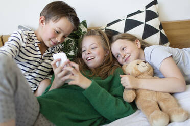 Group picture of three children lying together on bed looking at cell phone - HMEF00935
