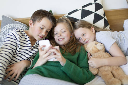 Gruppenbild von drei Kindern, die zusammen auf einem Bett liegen und auf ein Mobiltelefon schauen - HMEF00936