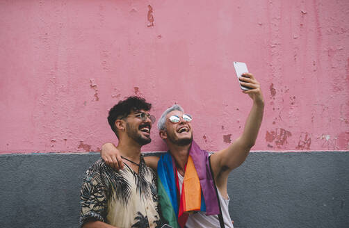 Gay couple taking a selfie in front of pink and grey wall - JCMF00724