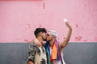 Gay couple kissing and taking a selfie in front of pink and grey wall - JCMF00725