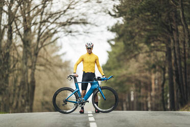 Lächelnder junger Mann in Sportkleidung steht mit Mountainbike auf einer Landstraße - MTBF00396