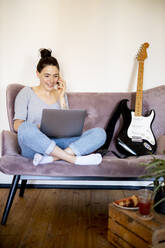 Porträt einer lächelnden jungen Frau am Telefon, die zu Hause auf der Couch sitzt und auf den Laptop schaut - DAWF01453