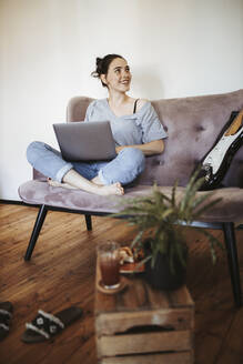 Porträt einer lächelnden jungen Frau, die zu Hause auf der Couch mit einem Laptop sitzt - DAWF01457