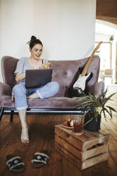 Portrait of smiling young woman sitting on couch at home using smartphone - DAWF01460
