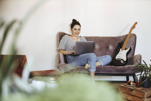 Portrait of smiling young woman sitting on couch at home using laptop - DAWF01461