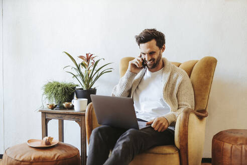 Smiling man on the phone sitting on armchair at home looking at laptop - DAWF01469